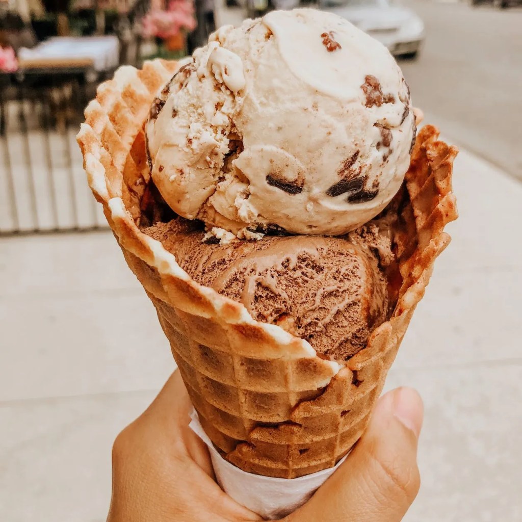 Schoko-Kugel Eis im Hörnchen, die in einer Hand getragen wird.