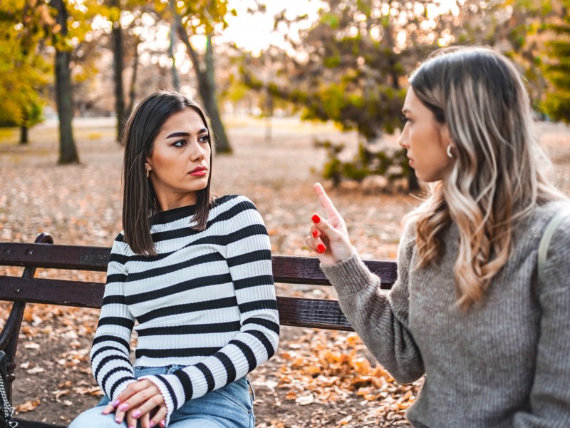 Zwei Frauen auf einer Parkbank im Herbst, die sich gegenseitig beschuldigen und ernst anschauen.c
