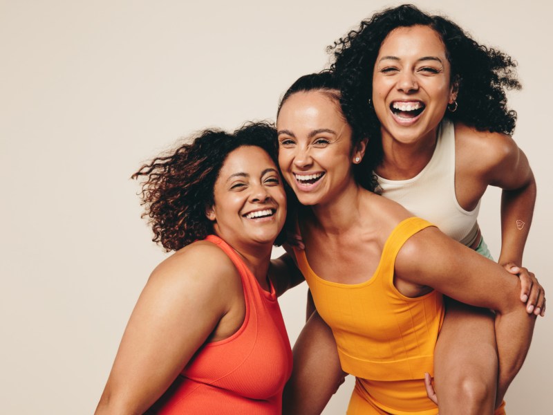 Drei Frauen in Sportklamotten, die zusammen lachen und vor einer hellbraunen Wand in die Kamera lächeln.