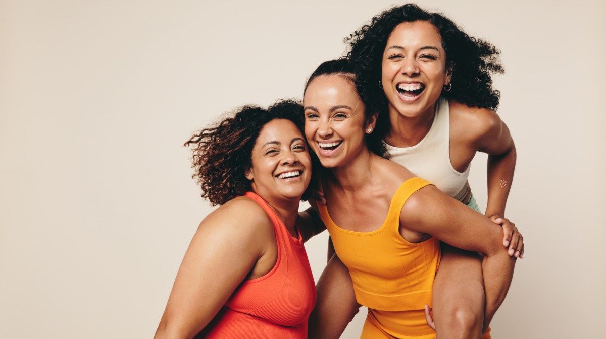 Drei Frauen in Sportklamotten, die zusammen lachen und vor einer hellbraunen Wand in die Kamera lächeln.