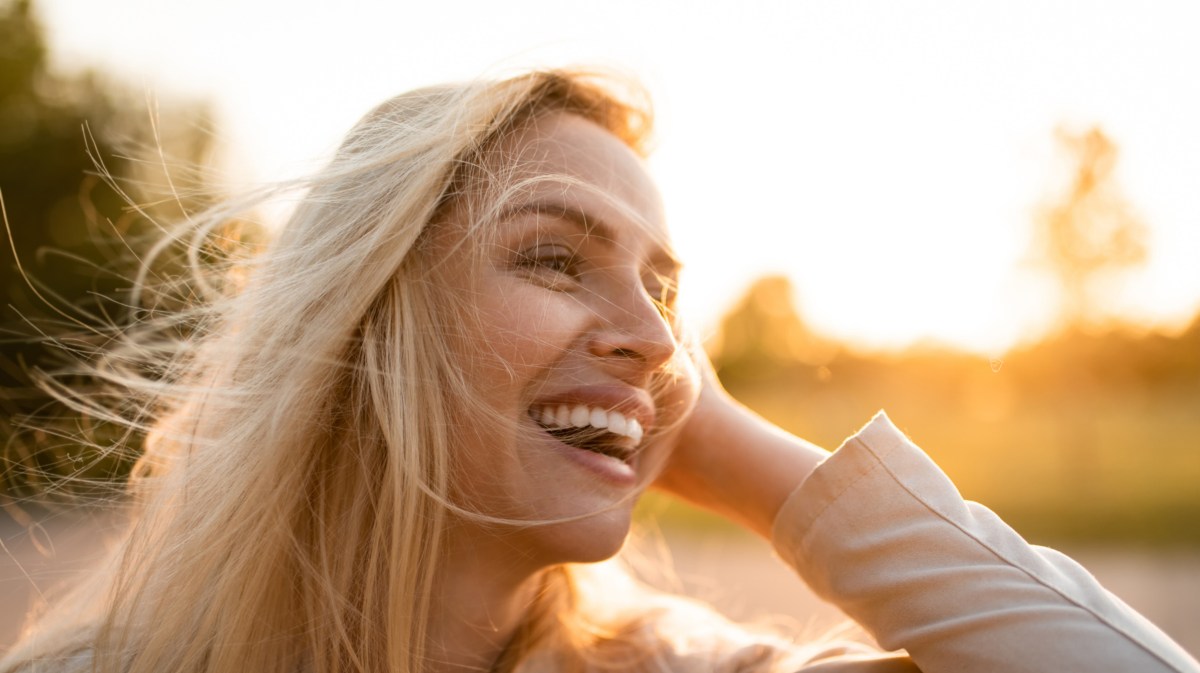 Frau mit blonden Haaren, die zur Seite lächelt und ihre Haare wegstreicht.