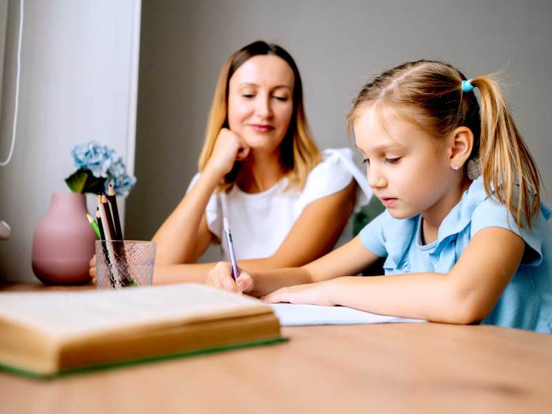 Mutter sitzt neben ihrer Tochter am Schreibtisch und schaut genau, wie das Kind seine Hausaufgaben macht.