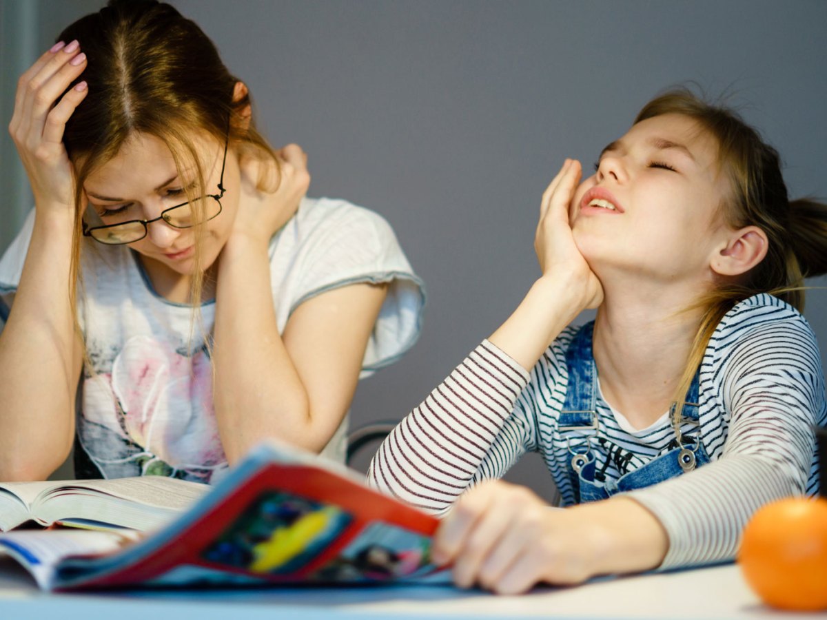 Mutter und Tochter sitzen gemeinsam am Tisch. Die Mutter liest etwas, die Tochter schaut verzweifelt aus einem Heft nach oben.