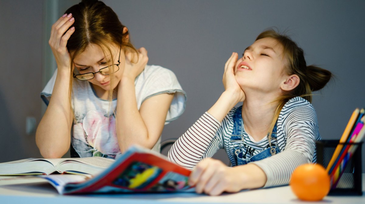 Mutter und Tochter sitzen gemeinsam am Tisch. Die Mutter liest etwas, die Tochter schaut verzweifelt aus einem Heft nach oben.