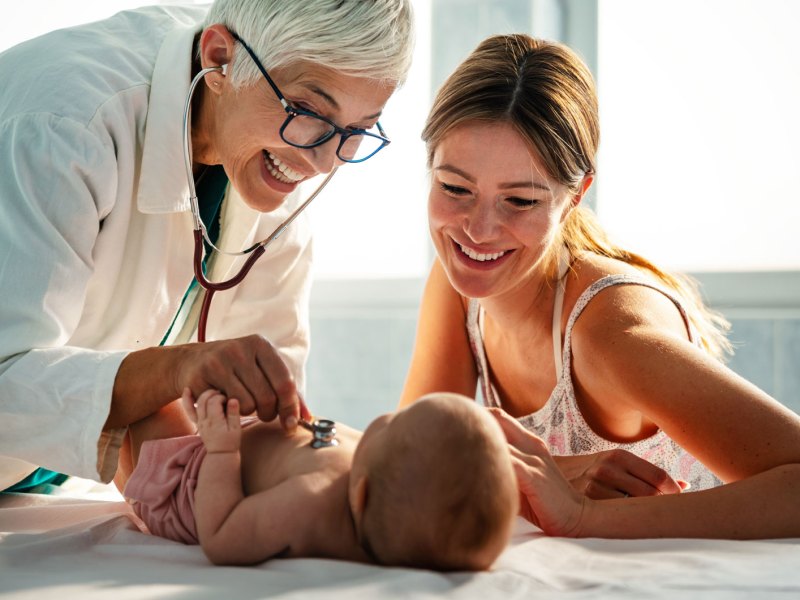 Ärztin untersucht ein kleines Baby, das auf der Liege liegt. Neben ihm sitzt die Mutter und strahlt es an.