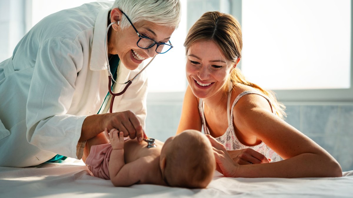 Ärztin untersucht ein kleines Baby, das auf der Liege liegt. Neben ihm sitzt die Mutter und strahlt es an.