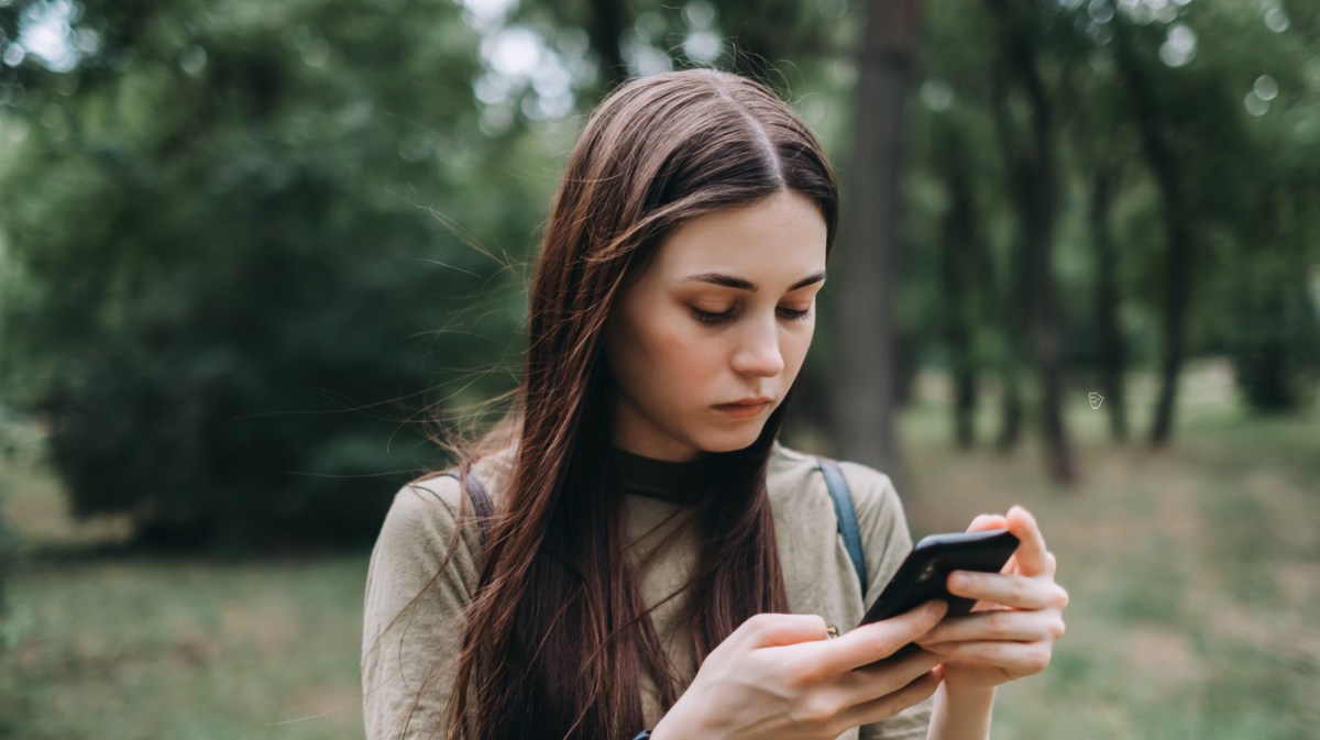 Frau auf einer Wiese mit Bäumen im Hintergrund, die nachdenklich auf ihr Handy runterschaut.