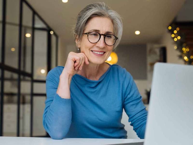 Frau mit blauem Shirt über 60 sitzt auf der Arbeit vor einem Laptop und lächelt.