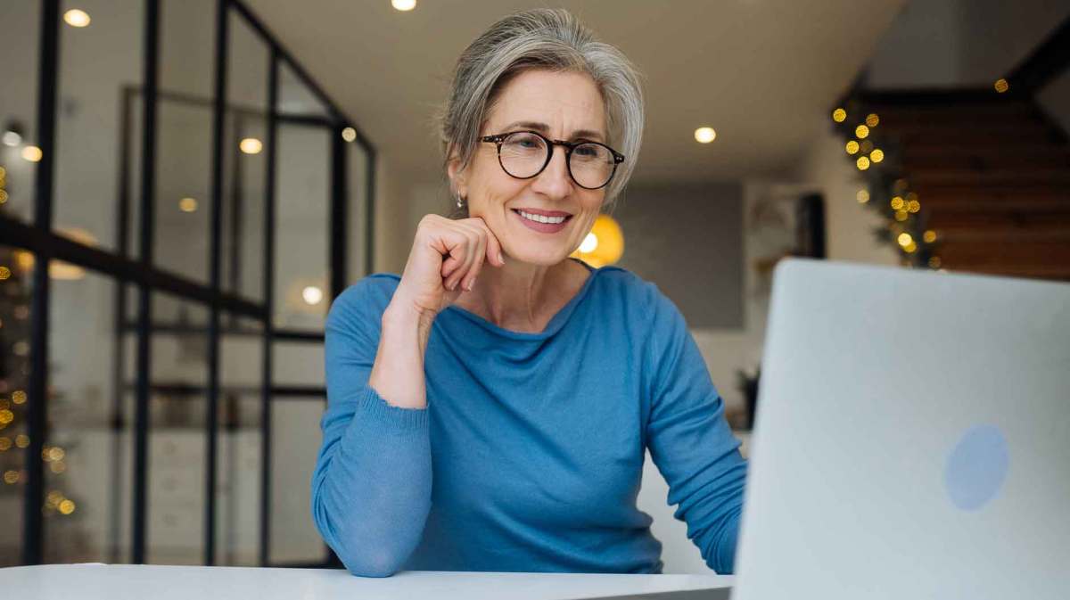 Frau mit blauem Shirt über 60 sitzt auf der Arbeit vor einem Laptop und lächelt.