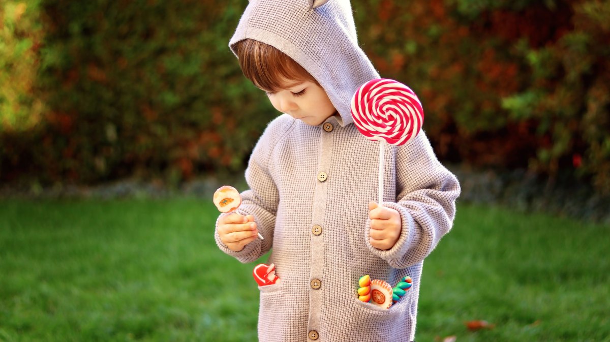 Ein Kind, ca. 4 oder 5 Jahre alt, steht draußen, mit einem großen Lolli in der Hand und weiteren kleinen in den Taschen seiner Strickjacke.