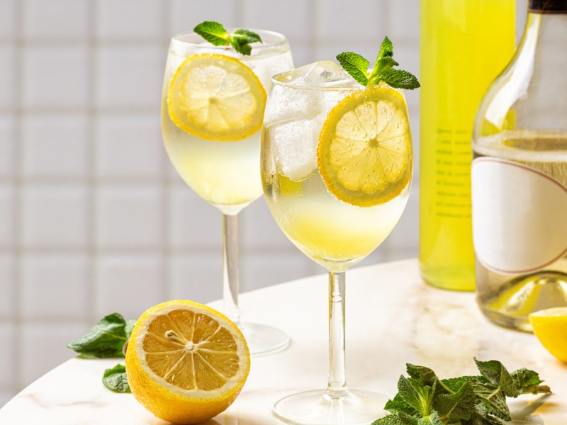 Zwei Gläser mit Limoncello Spritz auf einem weißen Tisch, eine halb aufgeschnittene Zitrone daneben.