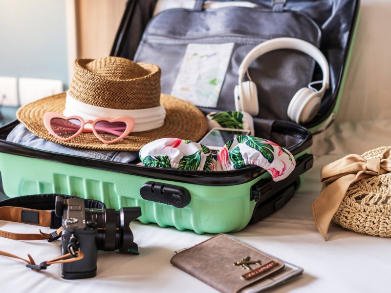 Grüner geöffneter Koffer auf Bett, in dem mehrere Dinge liegen, wie Sonnenhut, Kopfhörer.