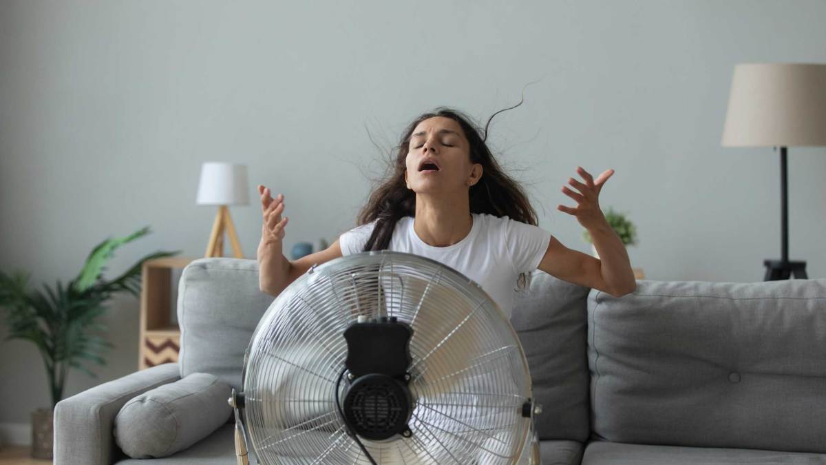 Frau sitzt auf der Couch im Wohnzimmer und atmet frische Luft aus dem Bodenventilator, weil sie keine Klimaanlage hat. Millennial-Frau benutzt Ventilator und kämpft mit der Hitze zu Hause.