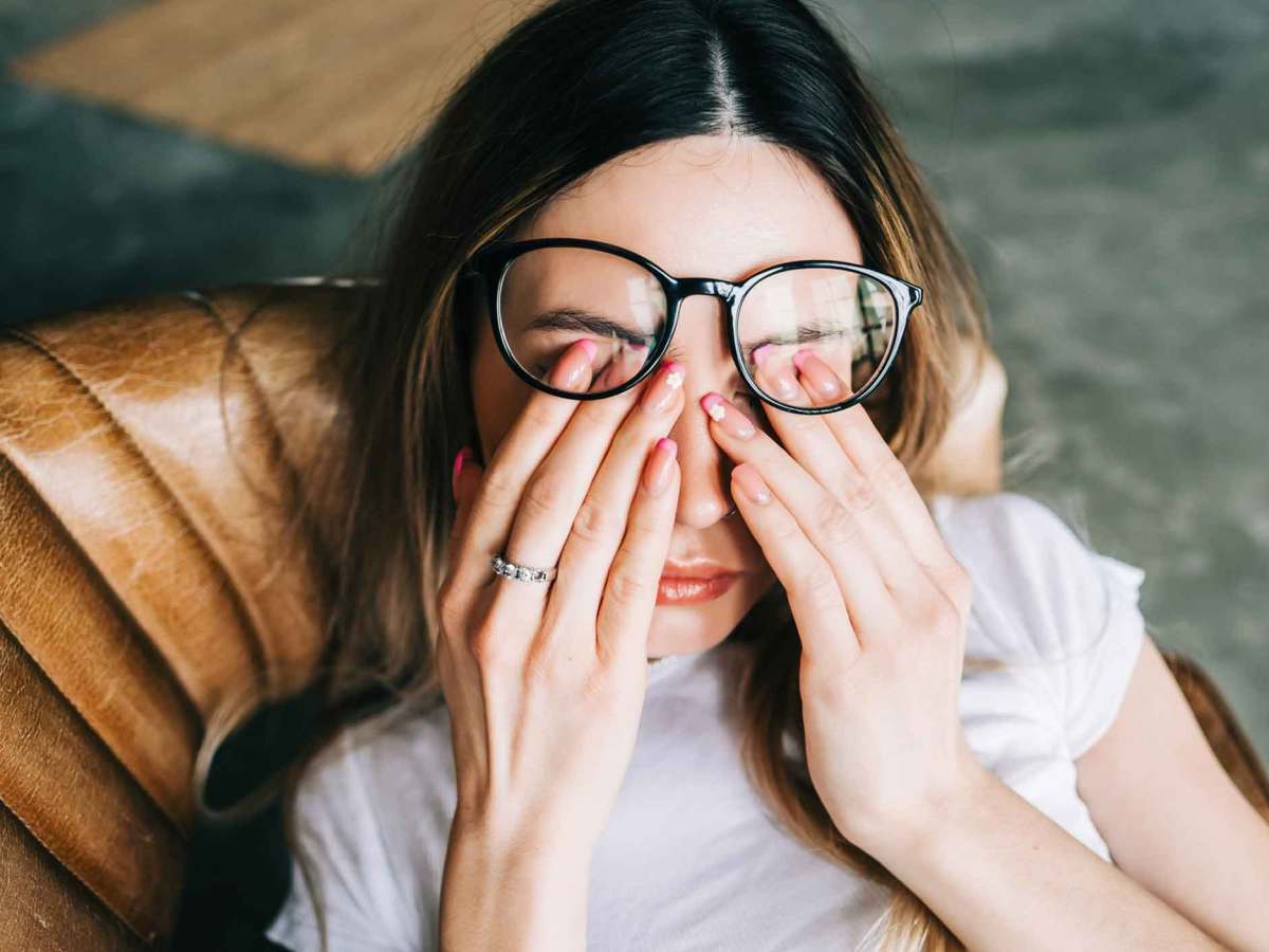 Frau sitzt auf einem Stuhl und reibt sich die Augen. Dabei rutscht ihre Brille nach oben.