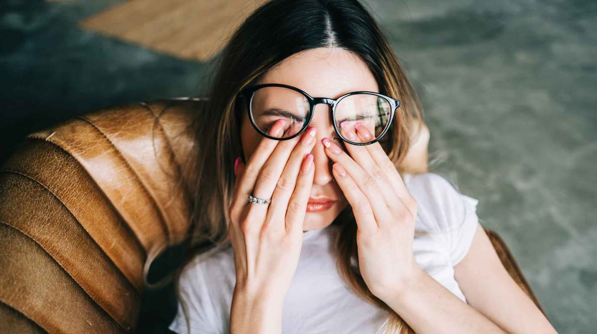 Frau sitzt auf einem Stuhl und reibt sich die Augen. Dabei rutscht ihre Brille nach oben.