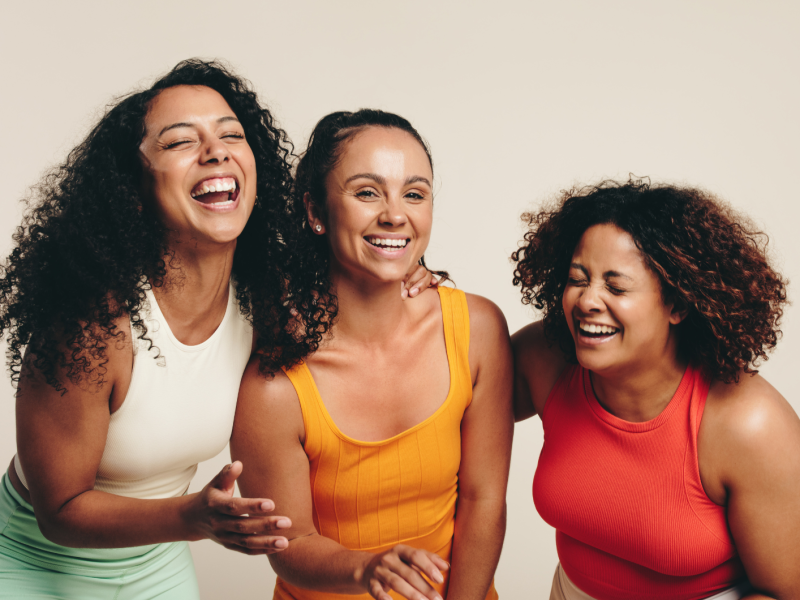 Drei Frauen in Sportklamotten vor einer beigen Wand, die gemeinsam lachen.