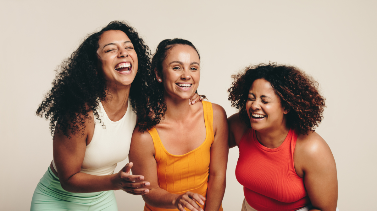 Drei Frauen in Sportklamotten vor einer beigen Wand, die gemeinsam lachen.