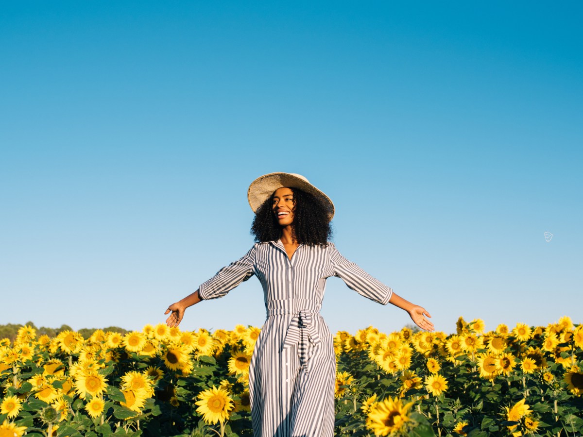 Frau mit Hut und Kleid in einem Sonnenblumenfeld die ihre Arme zur Seite ausbreitet und lacht.