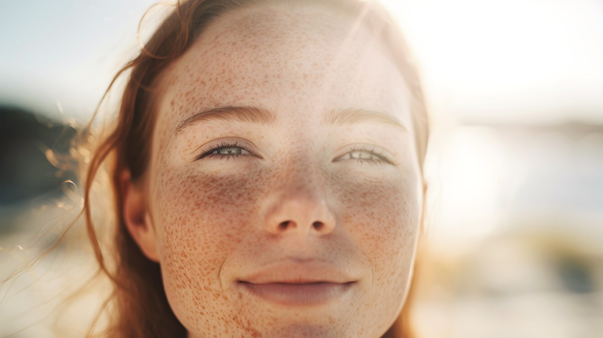 Frau mit roten Haaren am Strand, die von Sonnenstrahlen angeleuchtet wird und in die Kamera lächelt.