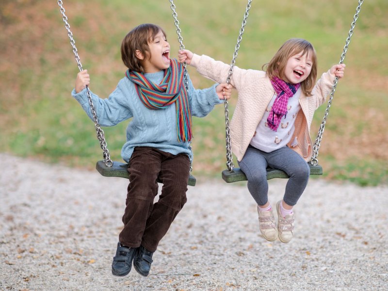 Zwei Kinder, ein Junge und ein etwas jüngeres Mädchen, sitzen draußen auf einer Schaukel und albern herum.