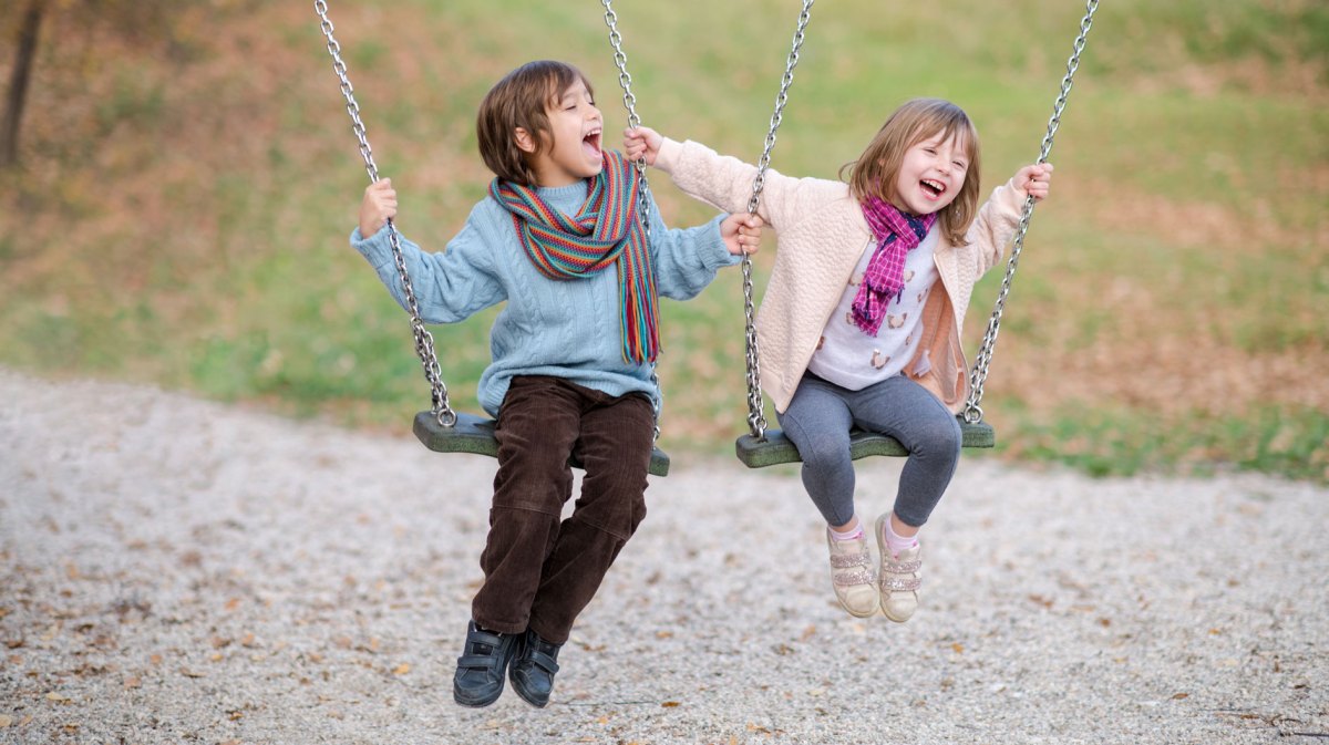 Zwei Kinder, ein Junge und ein etwas jüngeres Mädchen, sitzen draußen auf einer Schaukel und albern herum.