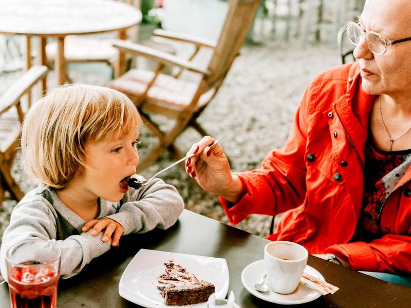 Großmutter füttert ihrem ca. 4-jährigen Enkel ein Stück Kuchen in einem Straßencafé.