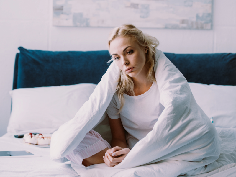 Frau alleine im Bett, mit einer Decke umschlungen, die gelangweilt nach vorne schaut.