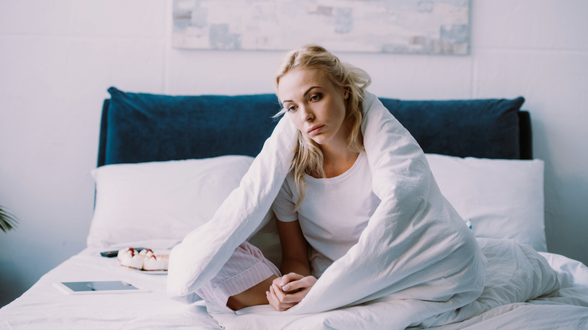 Frau alleine im Bett, mit einer Decke umschlungen, die gelangweilt nach vorne schaut.