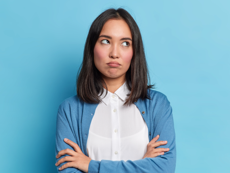 Frau vor einer blauen Wand, die genervt nach oben blickt und die Arme verschränkt.