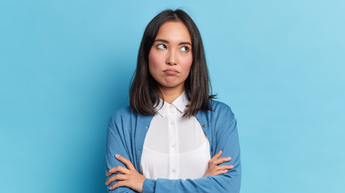 Frau vor einer blauen Wand, die genervt nach oben blickt und die Arme verschränkt.