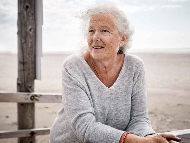 Ältere Frau steht am Strand an einen Zaun angelehnt.