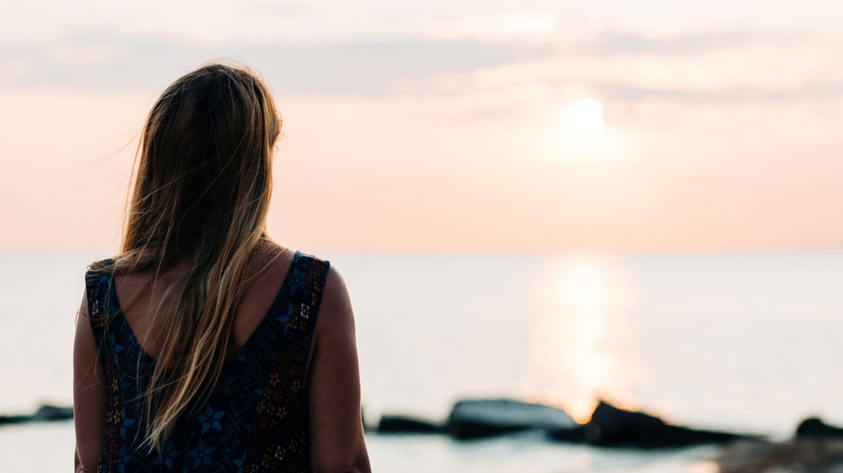 Frau steht am Meer und schaut den Sonnenuntergang