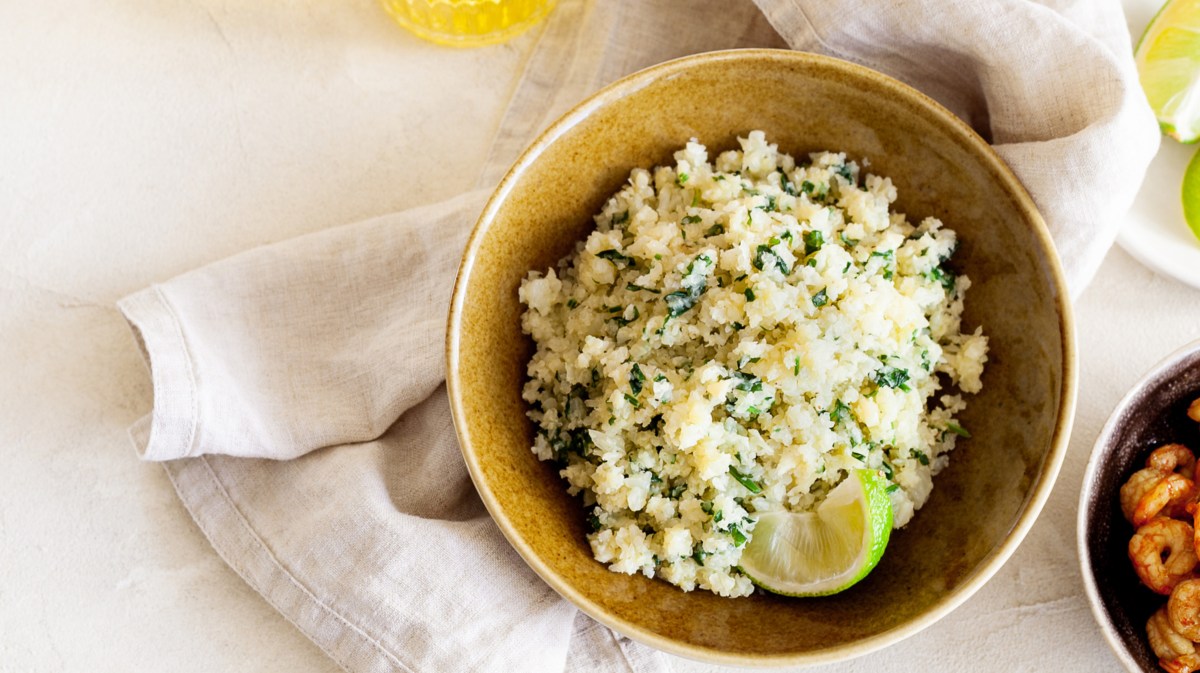 Blumenkohlreis mit Feta und Gewürzen in einer gelben Schüssel auf einem Esstisch, darunter liegt ein beiges Küchentuch.