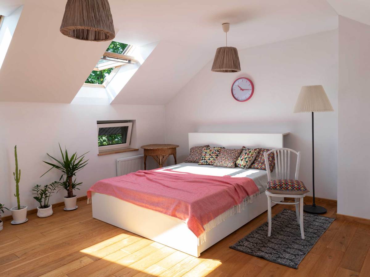 Helles Schlafzimmer mit Dachfenstern, weißem Bett, bunten Kissen, rosa Plaid und Topfpflanzen auf Holzfußboden.