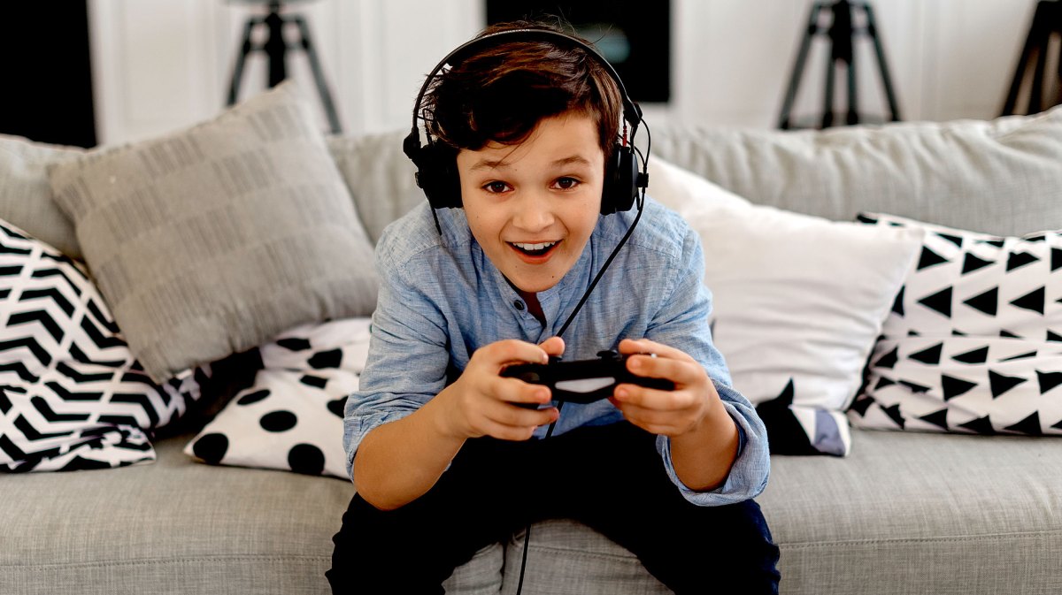 Junge, ca. 12 Jahre, sitzt mit Headset und Controller auf dem Sofa vor der Spielekonsole.