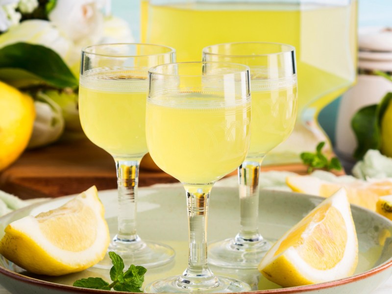 Drei kleine Gläser mit Limoncello stehen vor einer großen Flasche, umgeben von Zitronenspalten.