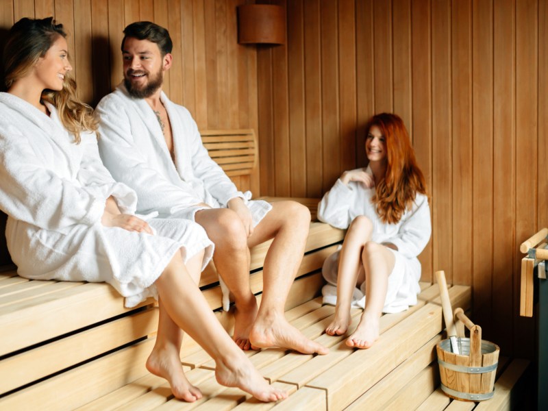 Drei Personen, ein Mann und zwei Frauen, tragen Bademäntel und sitzen in der Sauna.