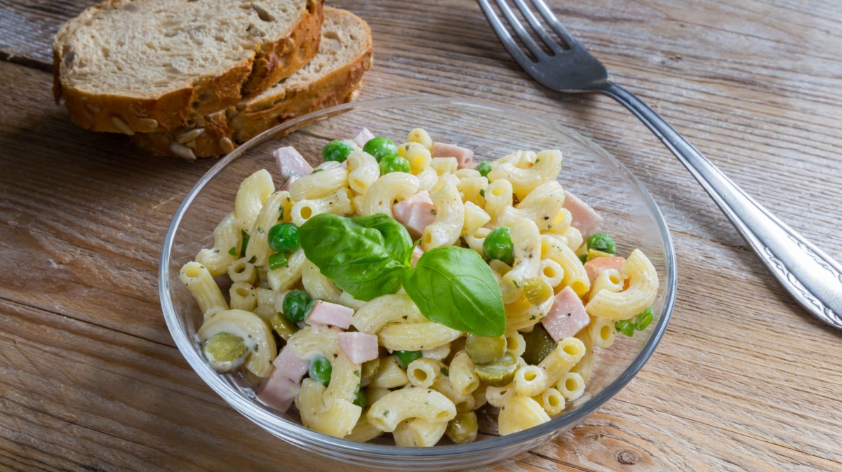 Nudelsalat in Glasschüssel mit Fleischwurst, Erbsen und Gurken.