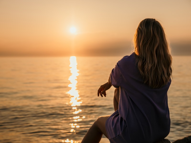 Frau sitzt am Meer und schaut sich Sonnenuntergang an.