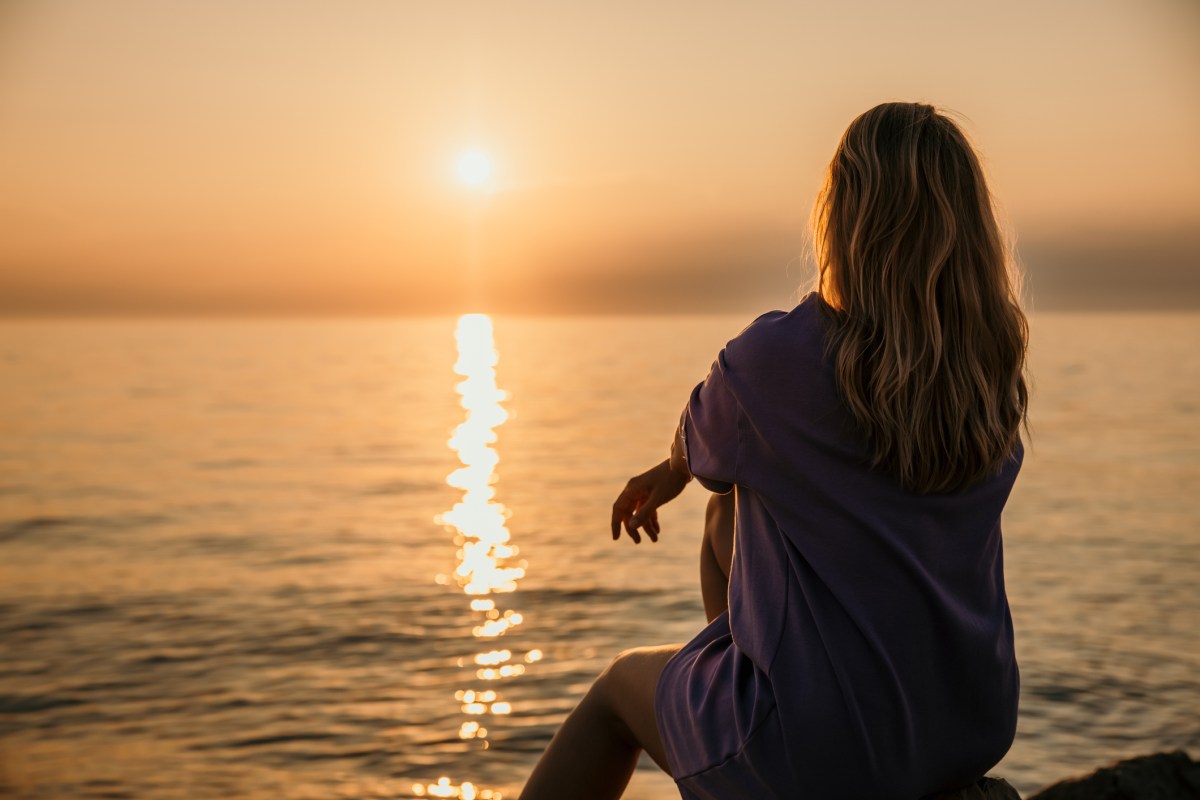 Frau sitzt am Meer und schaut sich Sonnenuntergang an.