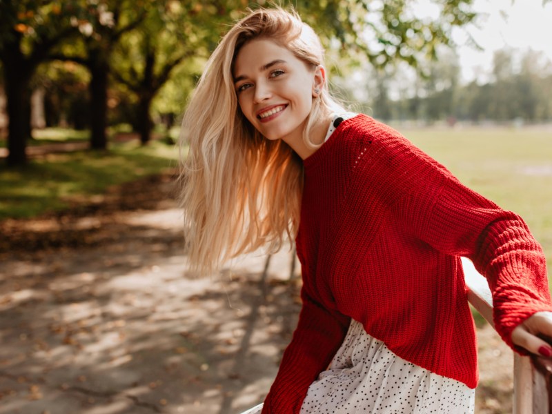 Junge Frau mit blonden Haaren und rotem Pulli schaut lächelnd in Kamera, draußem im Park.