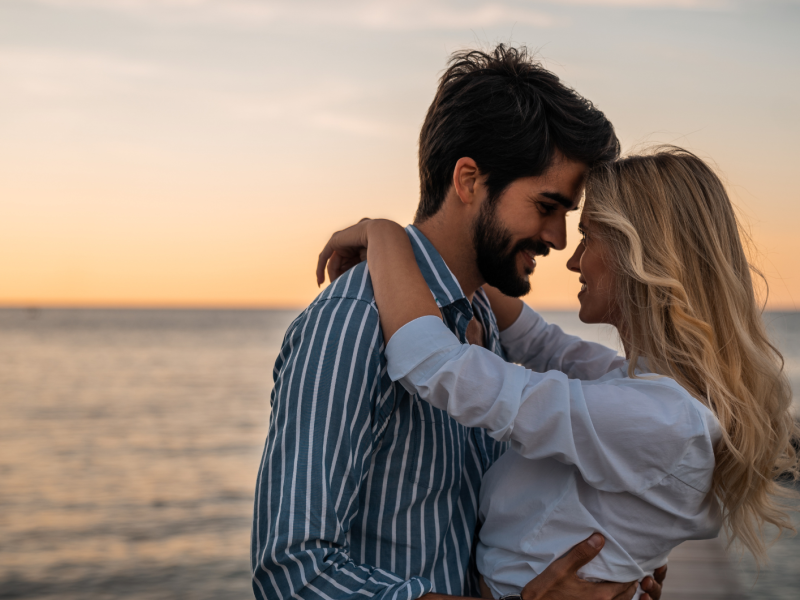 Frau und Manna am Strand beim Sonnenuntergang, die sich verliebt anschauen und umarmen.