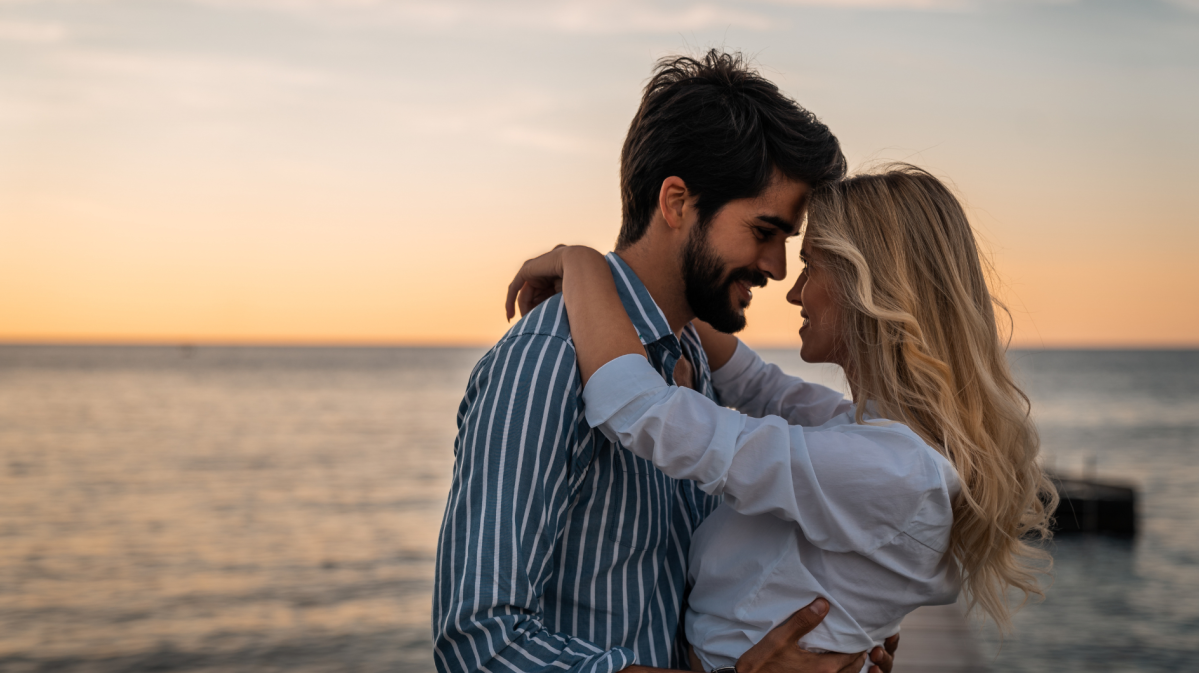 Frau und Manna am Strand beim Sonnenuntergang, die sich verliebt anschauen und umarmen.