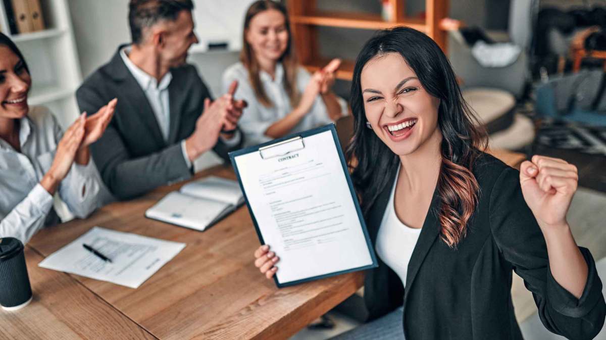 Junge Frau mit braunen Haaren sitzt auf der Arbeit und hält einen Vertrag hoch, über den sie sich sehr freut.