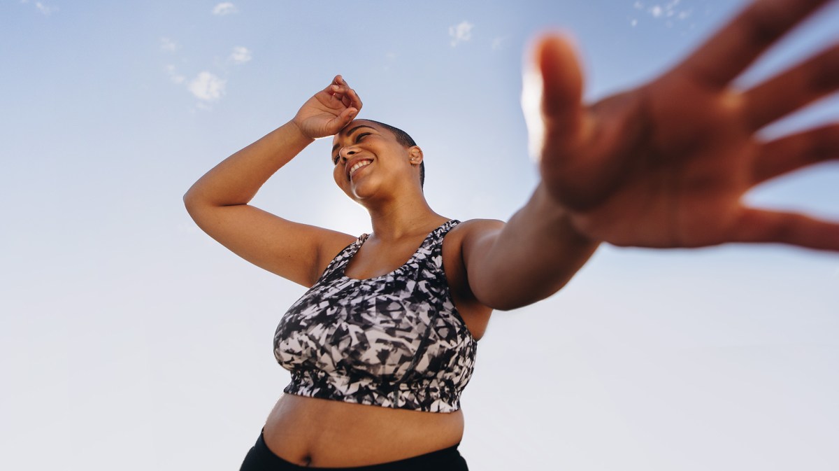 Junge plus-Size Frau in gemustertem Sport-BH lacht und streckt bei sonnigem Wetter die Hand in die Kamera vor einem klaren Himmel.