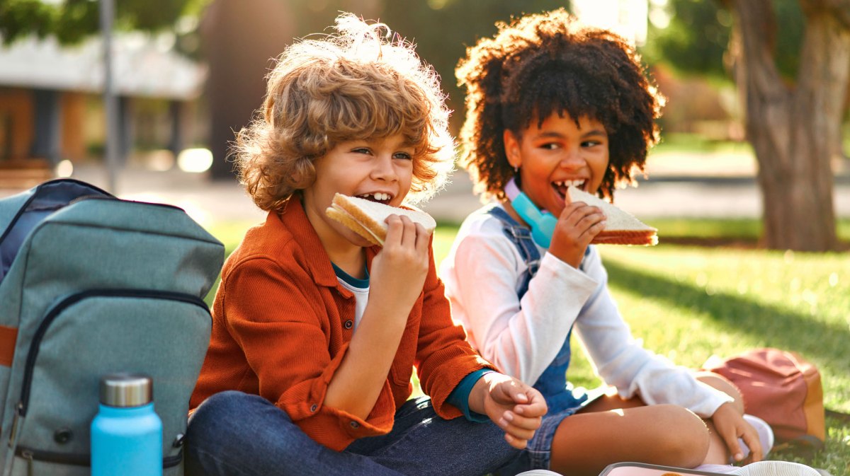 Zwei Schulkinder sitzen an einem sonnigen Tag auf der Wiese und essen ihre Schulbrote.