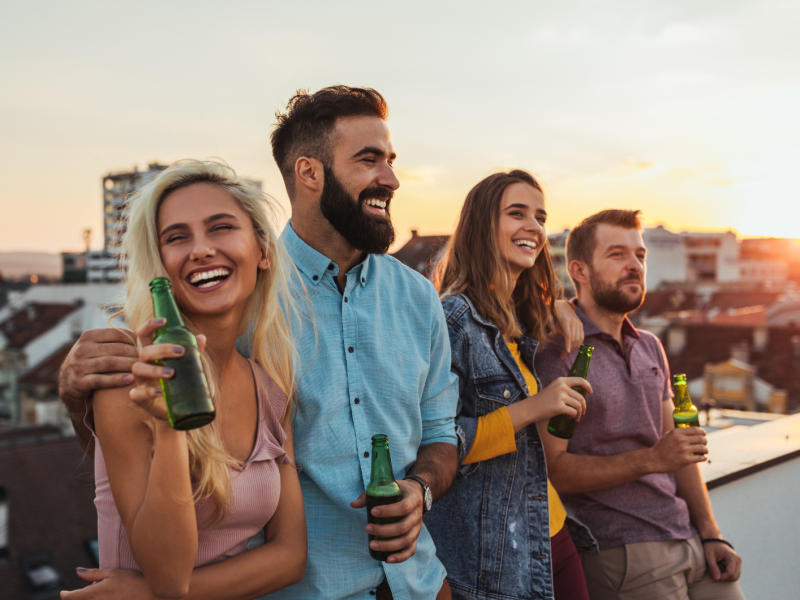 Zwei Pärchen auf einem Balkon, die nebeneinanderstehen, Bier in der Hand halten und lachen.