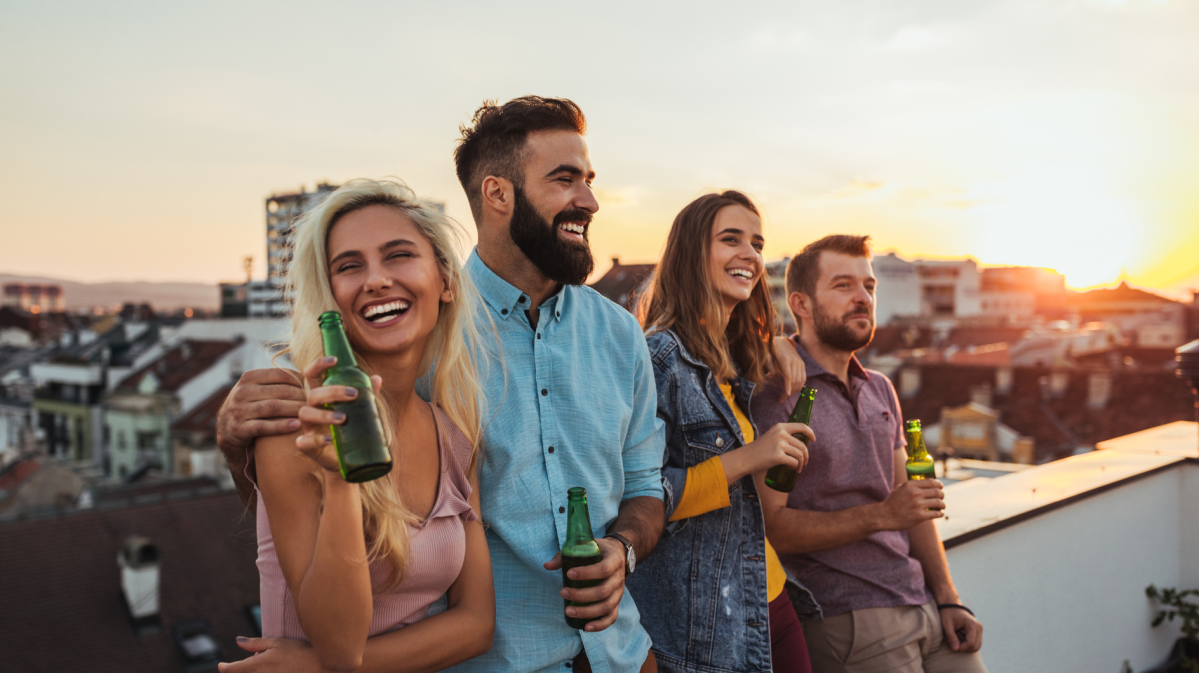 Zwei Pärchen auf einem Balkon, die nebeneinanderstehen, Bier in der Hand halten und lachen.