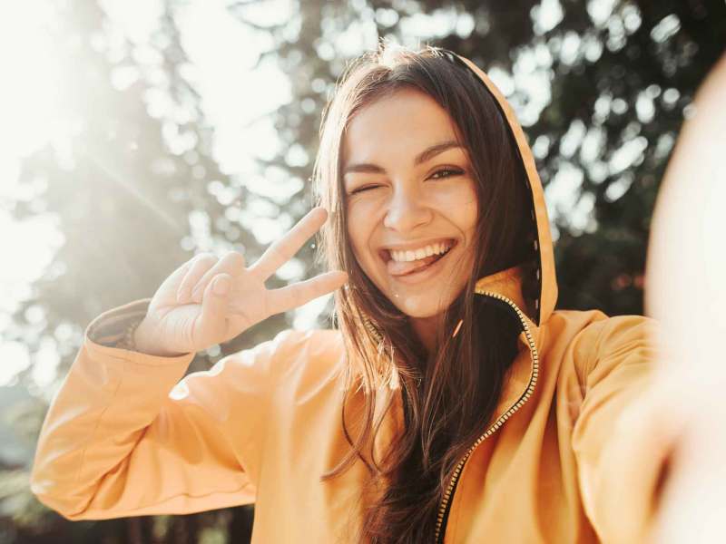 Junge Frau mit braunen Haaren hat eine gelbe Kapuzenjacke an und macht ein Selfie.