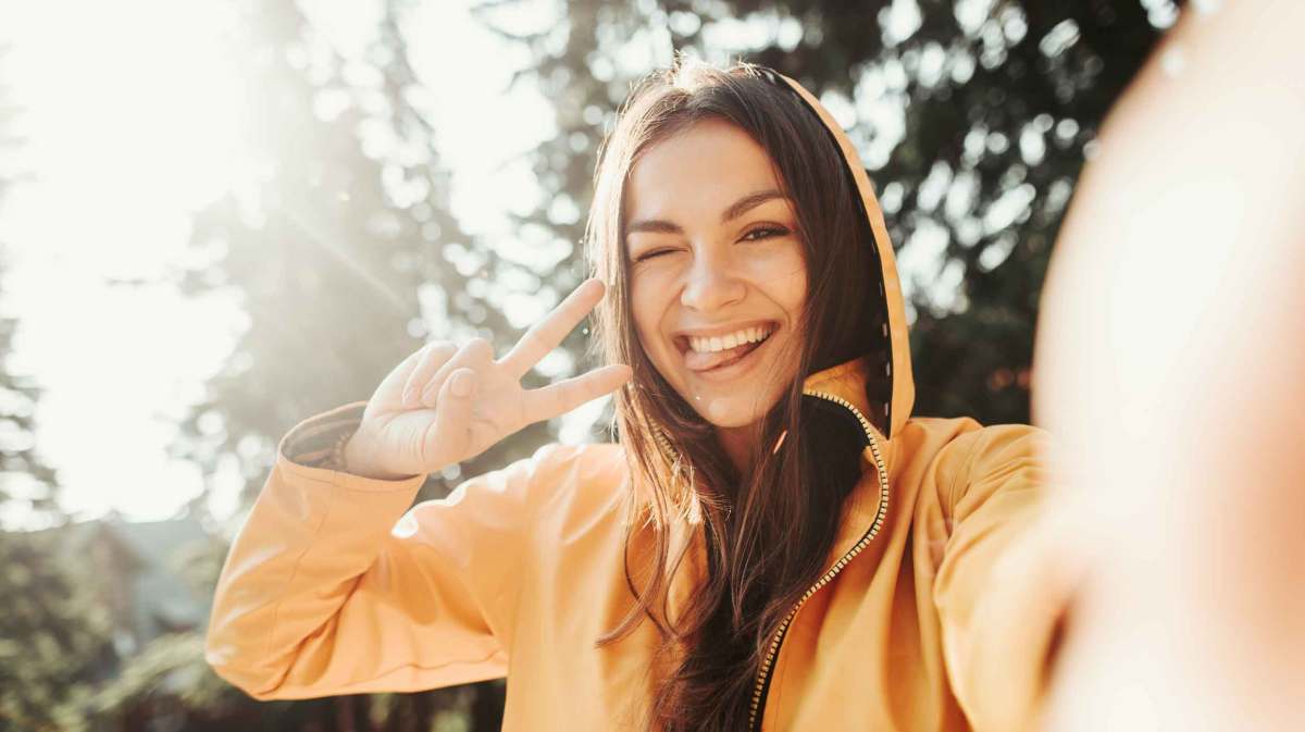 Junge Frau mit braunen Haaren hat eine gelbe Kapuzenjacke an und macht ein Selfie.